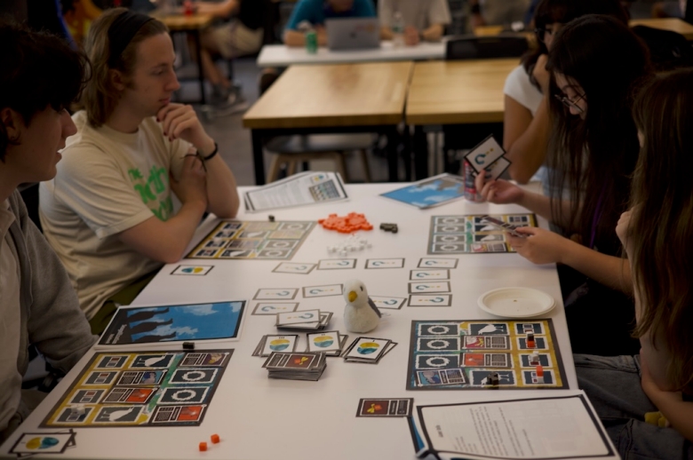 Group of Tisch Summer High school Game Design students sit at a table looking at cards with question marks and game mats during a game design class.